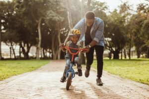 A man helping his son ride a bike on a park path surrounded by trees.