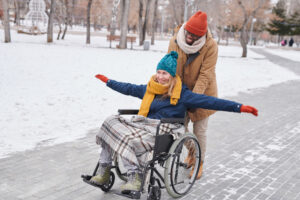 A smiling man pushing a smiling woman in a wheelchair at a park during winter to reduce the winter blues.