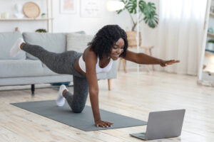 A woman doing a yoga pose in front of a laptop in a living room. Following along with online exercise videos is a great way to get active on a budget.