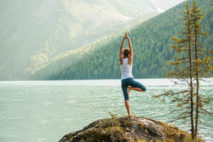 A woman practicing yoga on a rock in front of a lake surrounded by mountains.