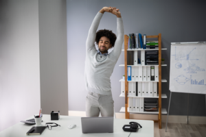 A man stretching his arms over his head while standing in front of a desk. 