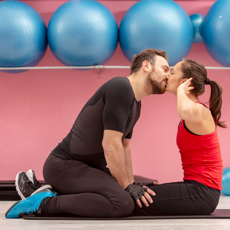A man kneels on his female partner’s legs to assist her with sit-ups. Large blue exercise balls appear on a rack in the background