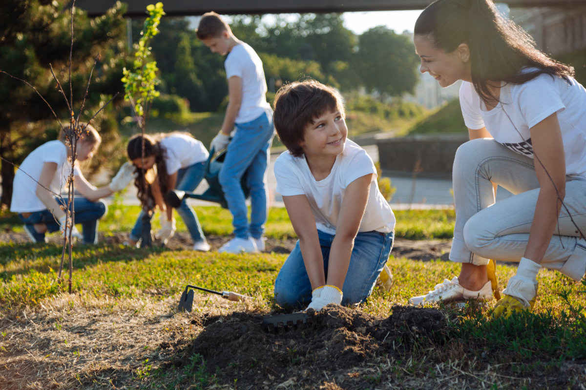 A kneeling woman  teaching a young boy how to garden while more youth are standing in the background also gardening