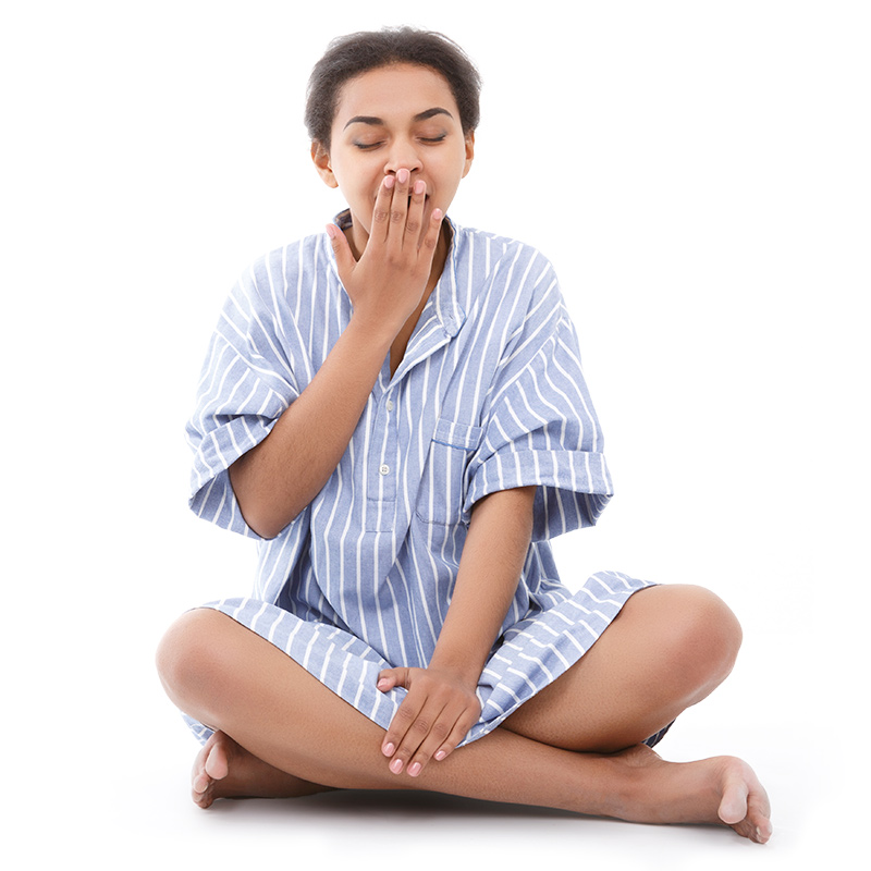 A young woman in pajamas sitting cross-legged and yawning 