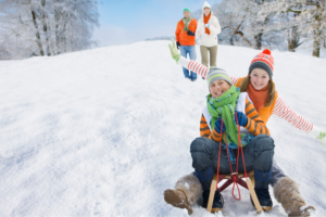 Two kids tobogganing down a hill while their parents walk behind them. 