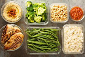 Hummus, broccoli, chickpeas, salsa, grilled chicken, green beans and rice in containers on a table. 