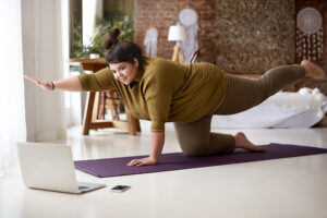 A woman stretching on a yoga mat while looking at a laptop. She is enjoying the benefits of fitness snacks.