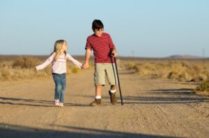 A girl holding hands with a boy walking with a cane on a dirt road. 