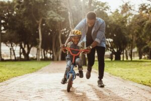 Un homme aide son fils à faire du vélo sur une piste de parc entourée d’arbres. 