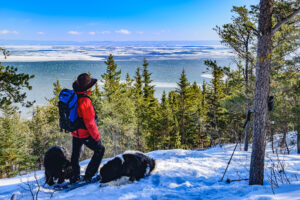 A person in hiking gear standing on a snow-covered hill overlooking trees and a body of water.