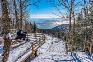 Two people sitting on a boardwalk overlooking a snow-covered hill, trees and a lake.