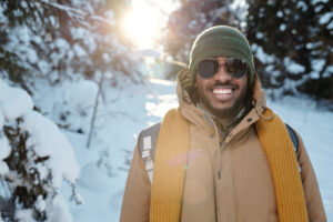A smiling man wearing a backpack and winter coat and standing on a snow-covered nature trail. 