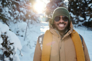 A smiling man wearing a backpack and winter coat and standing on a snow-covered nature trail.