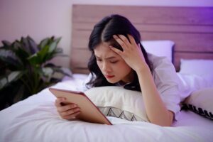 A worried-looking woman lying in bed while doomscrolling on a tablet.