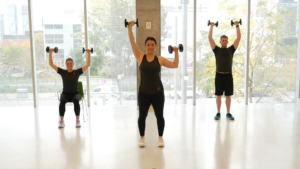 Three people holding pairs of dumbbells over their heads in a workout studio. 