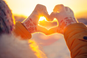 A person wearing winter gloves making a heart symbol with their hands while watching the sunset.