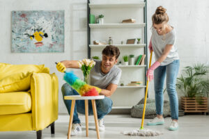 A man dusting a side table and a woman mopping a floor in a living room