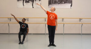 A person demonstrating ballet while seated in a chair beside a person demonstrating ballet standing up. 