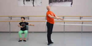 A person demonstrating ballet while seated in a chair beside a person demonstrating ballet standing up.