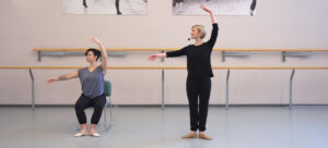 Une personne assise sur une chaise fait la démonstration d’une position de ballet à côté d’une personne qui exécute le même mouvement debout.