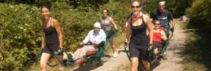Two people in wheelchairs being pulled by two pairs of people on a nature trail.