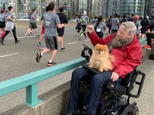 Sam Sullivan, un homme avec la tetraplégie, encourage des marathoniens alors qu’un chien est assis sur ses jambes. 