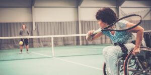 Une femme jouant au tennis en fauteuil roulant à l’intérieur contre un homme debout de l’autre côté du filet.