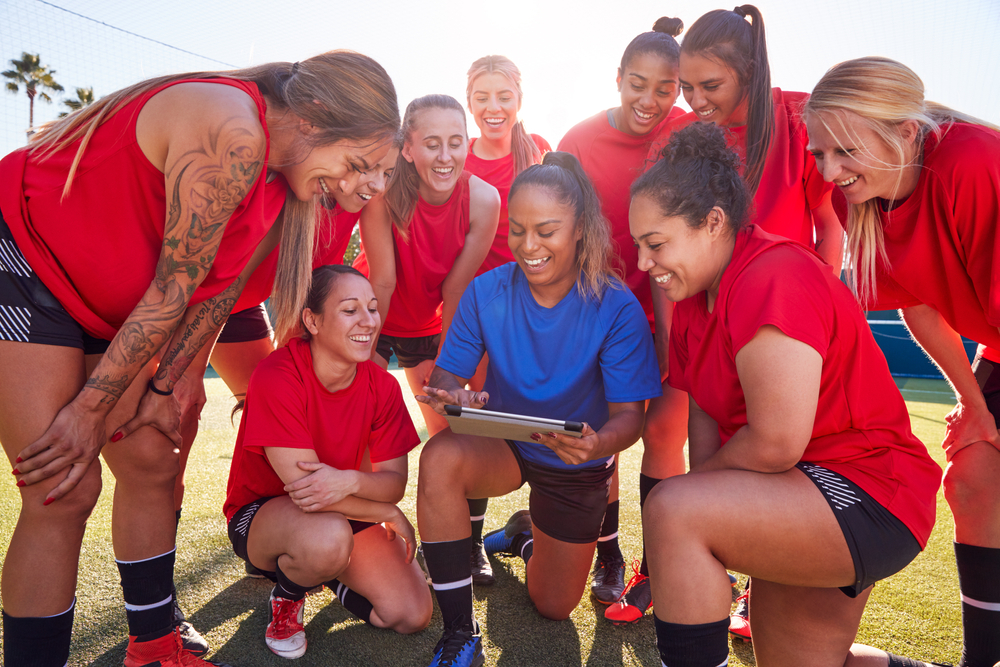 A group of women in red shirts looking at a tablet on a sports field.