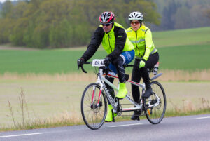 A man and woman riding a tandem bicycle on a road beside a field. 