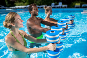 Three smiling people holding foam dumbbells in an outdoor pool.