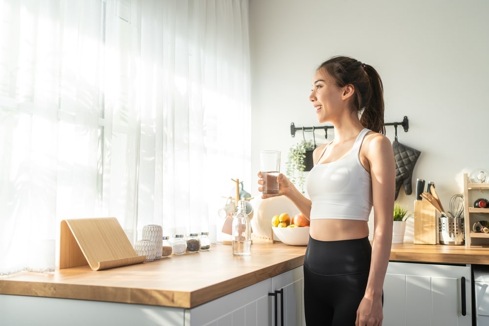 A smiling woman in workout clothes holding a glass of water in a kitchen. 