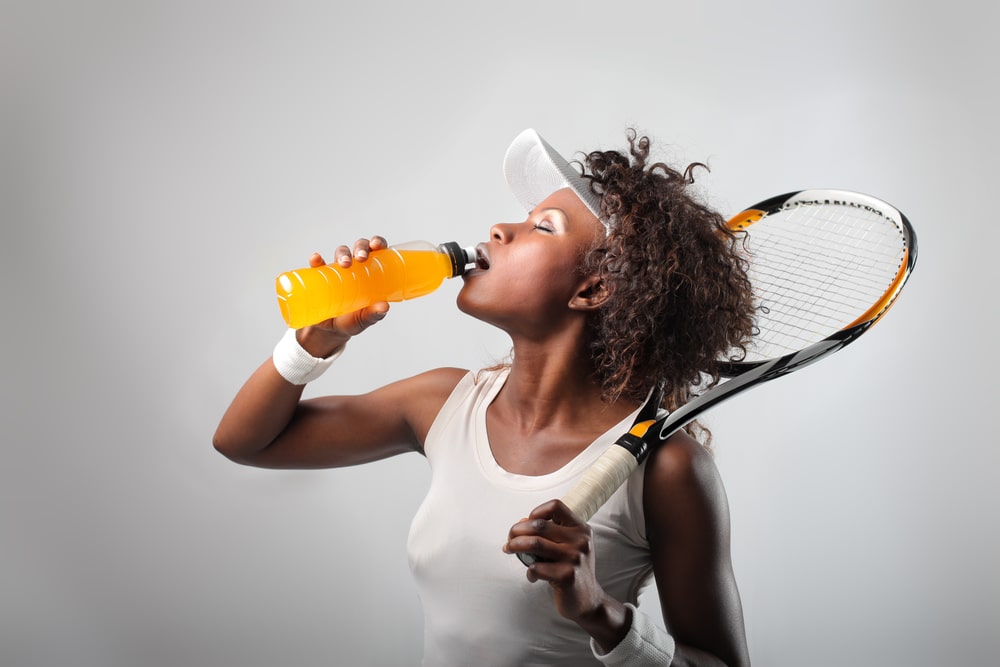 A woman drinking a sports drink while holding a tennis racquet 