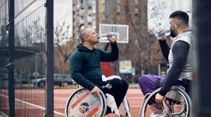 Two men in wheelchairs drinking water from bottles on an outdoor basketball court.