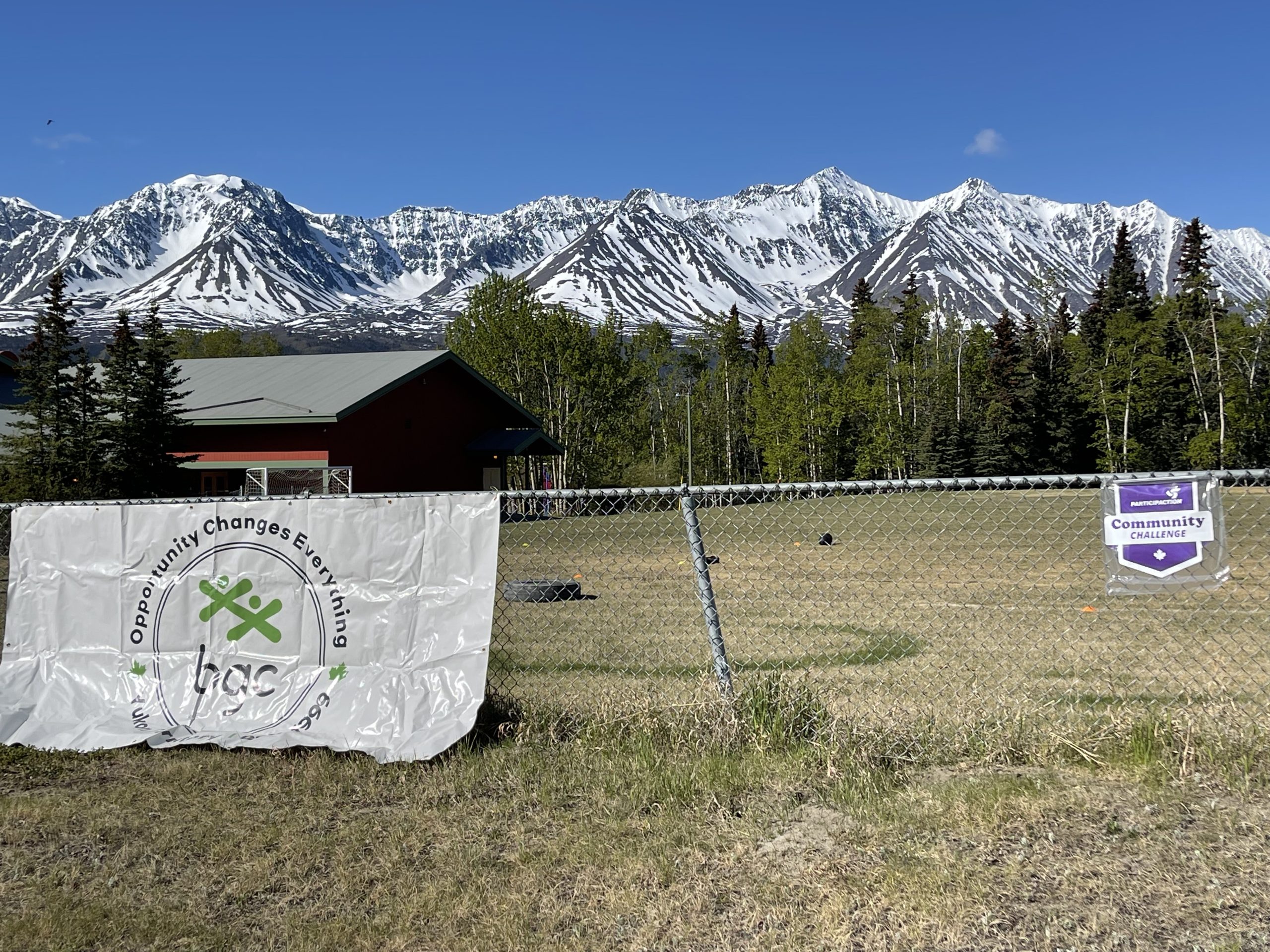 A fence with the BGC Yukon and ParticipACTION Community Challenge signs on it and a field, chalet, trees and mountains behind it. 