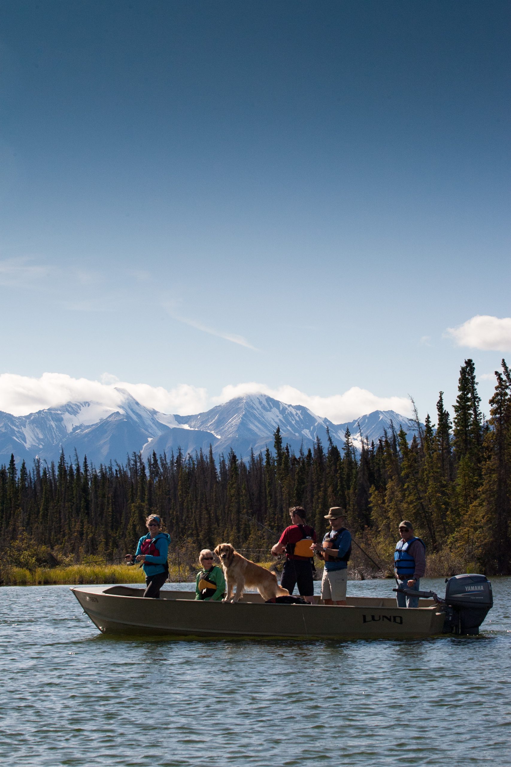 A dog and a group of people in a boat fishing on a lake with mountains and trees behind them. 