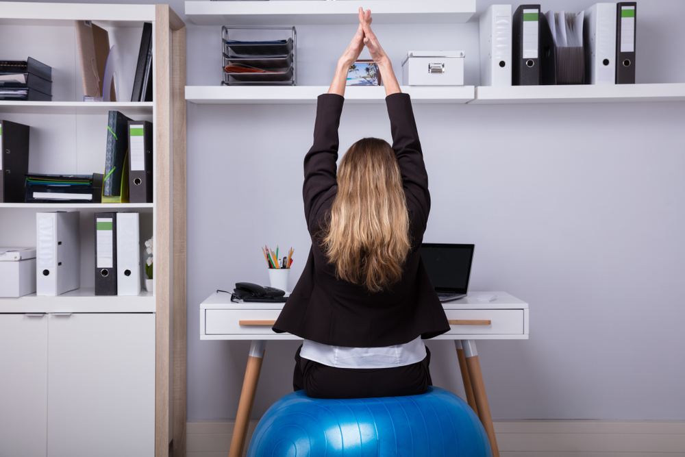 Une femme étire les bras au-dessus de la tête assise sur un ballon d’exercice devant un bureau.