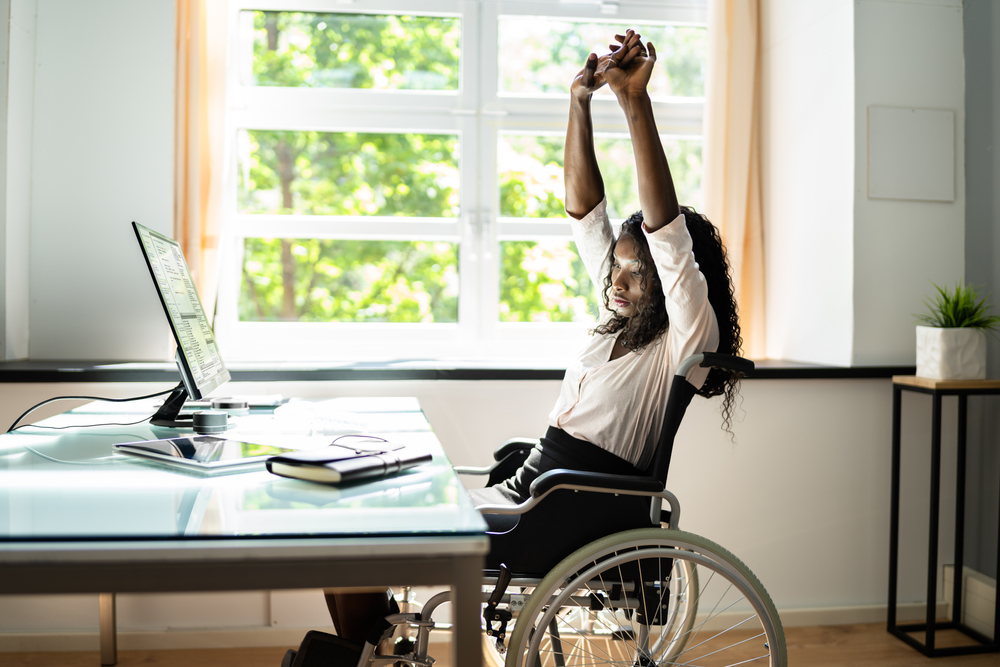 A woman in a wheelchair stretching her arms over her head while in front of a desk with a computer on it. 