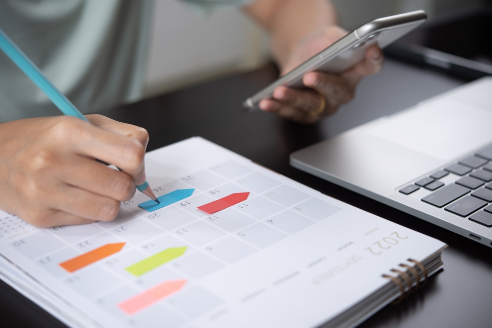 A person writing in a calendar while holding a phone in their other hand. 