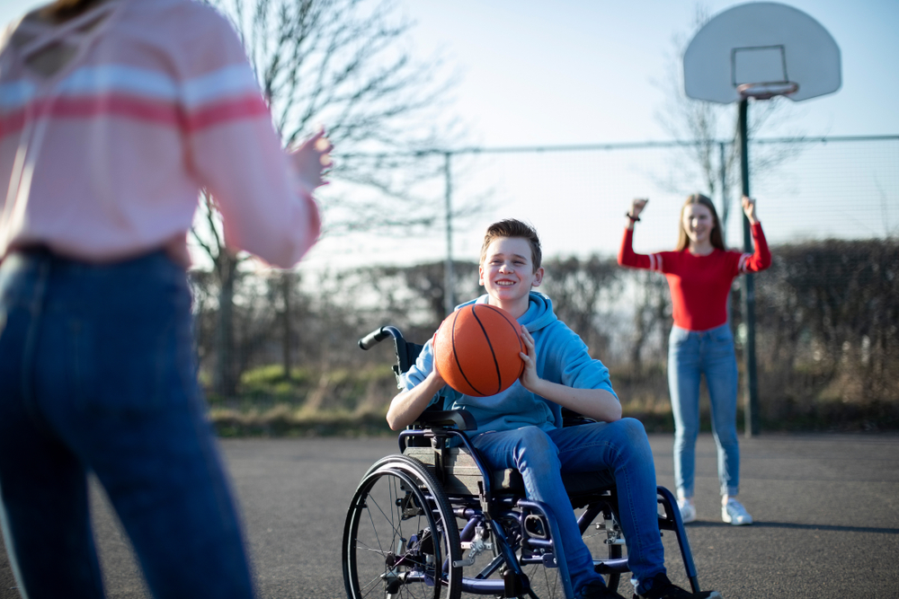 Un garçon dans un fauteuil roulant joue au basketball avec deux filles à l’extérieur. 