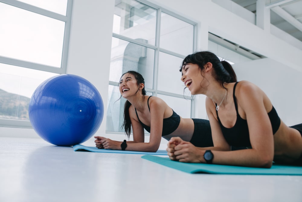 Two women laughing while laying on yoga mats beside an exercise ball.