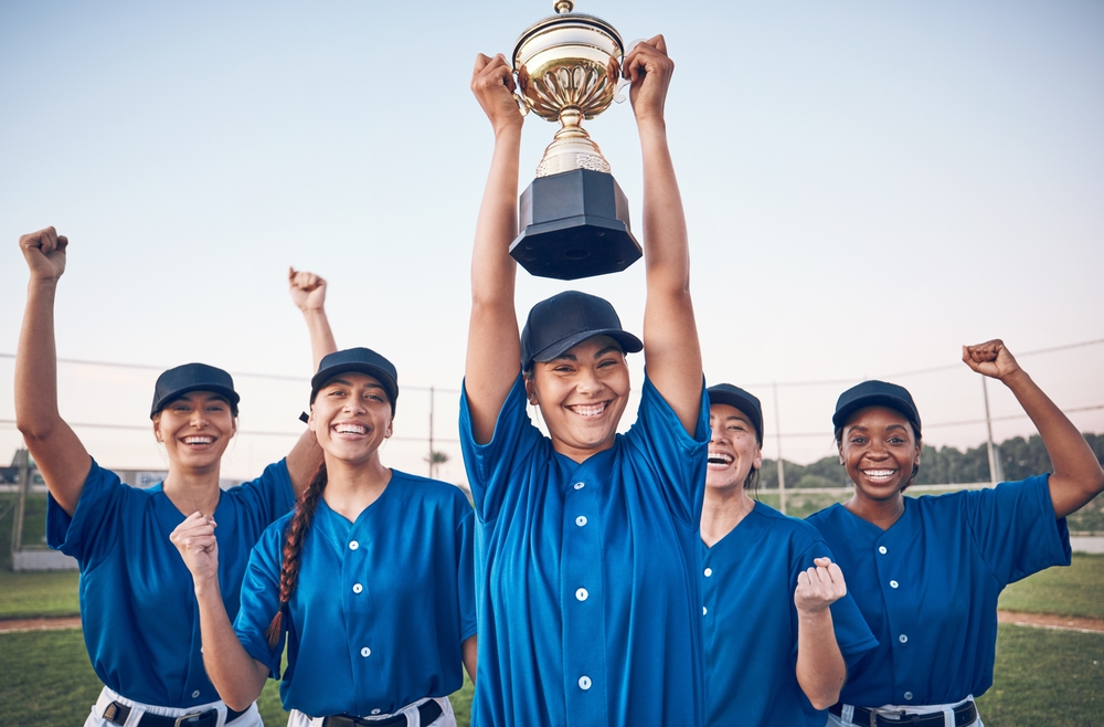 A female baseball player holding a trophy while her teammates cheer behind her. She's enjoying the mental health benefits of team sports. 