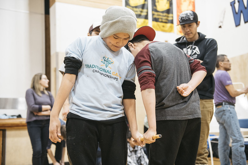 Two boys playing the Dene game called stick pull in a gymnasium. 