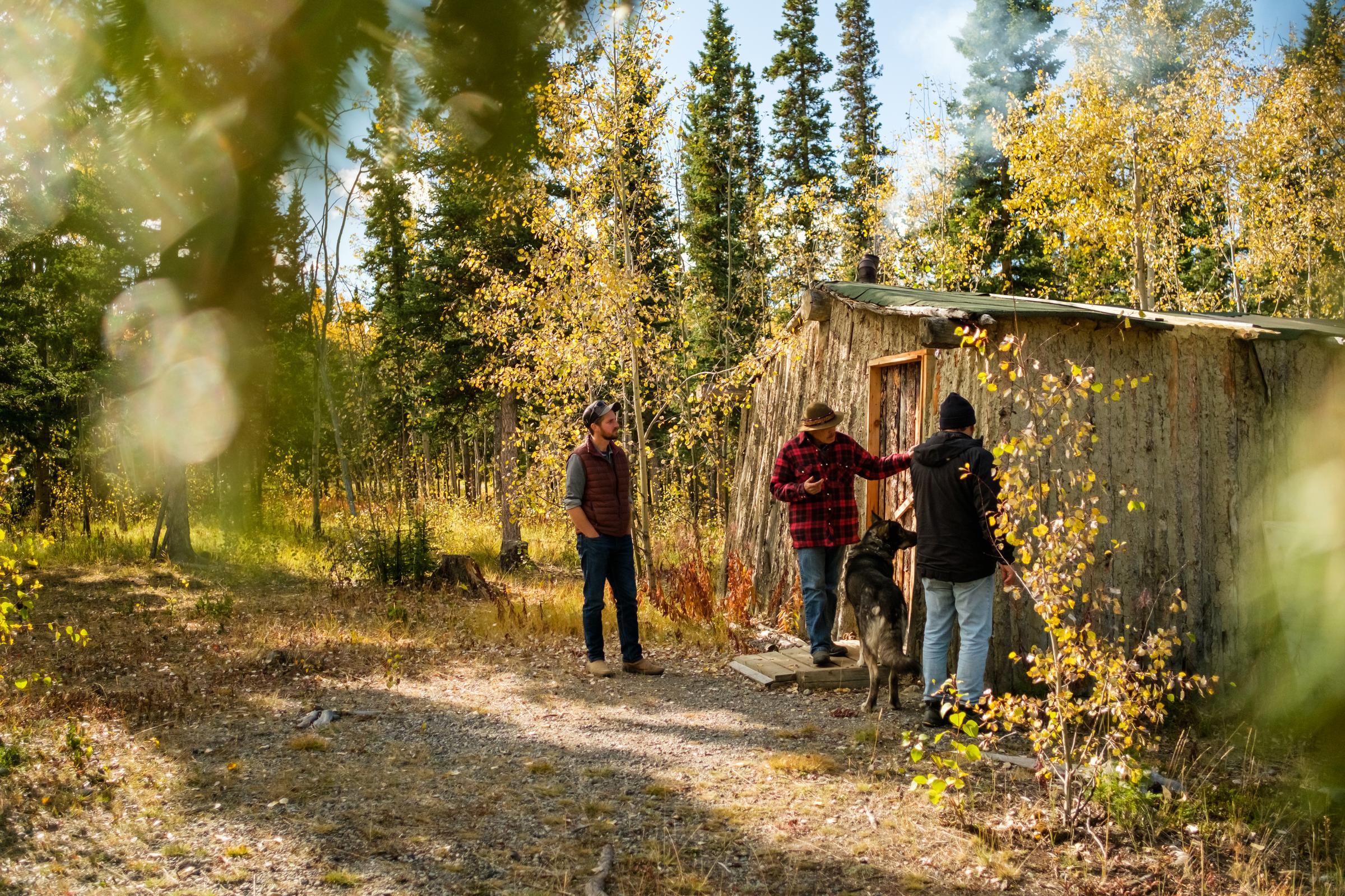 A group of men and a dog standing beside a log house in the woods. A group of men and a dog standing beside a log house in the woods.