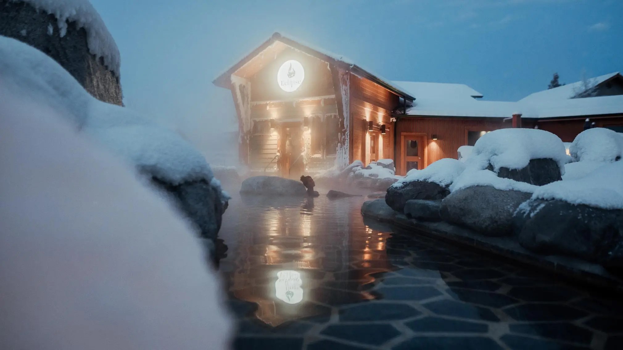 An outdoor hot pool in front of a snow-covered lodge. An outdoor hot pool in front of a snow-covered lodge.