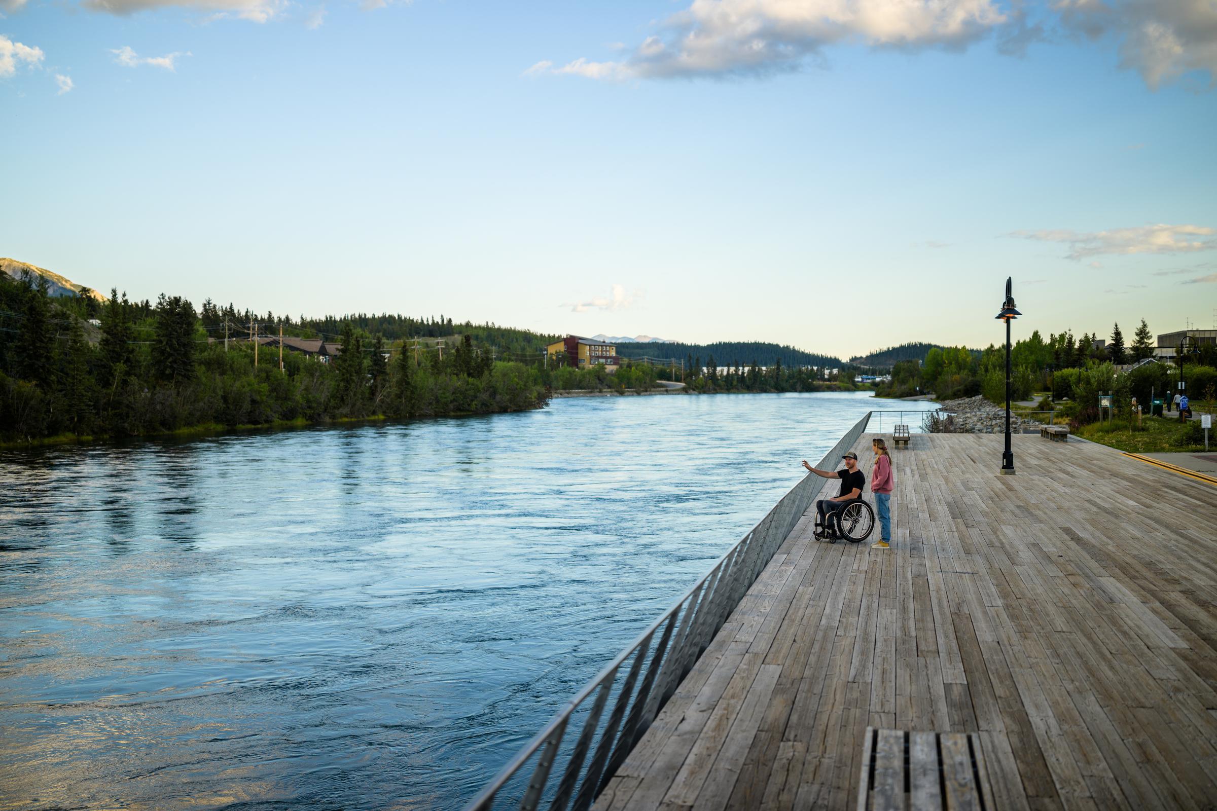 A man in a wheelchair and a woman on a boardwalk beside a river. A man in a wheelchair and a woman on a boardwalk beside a river.