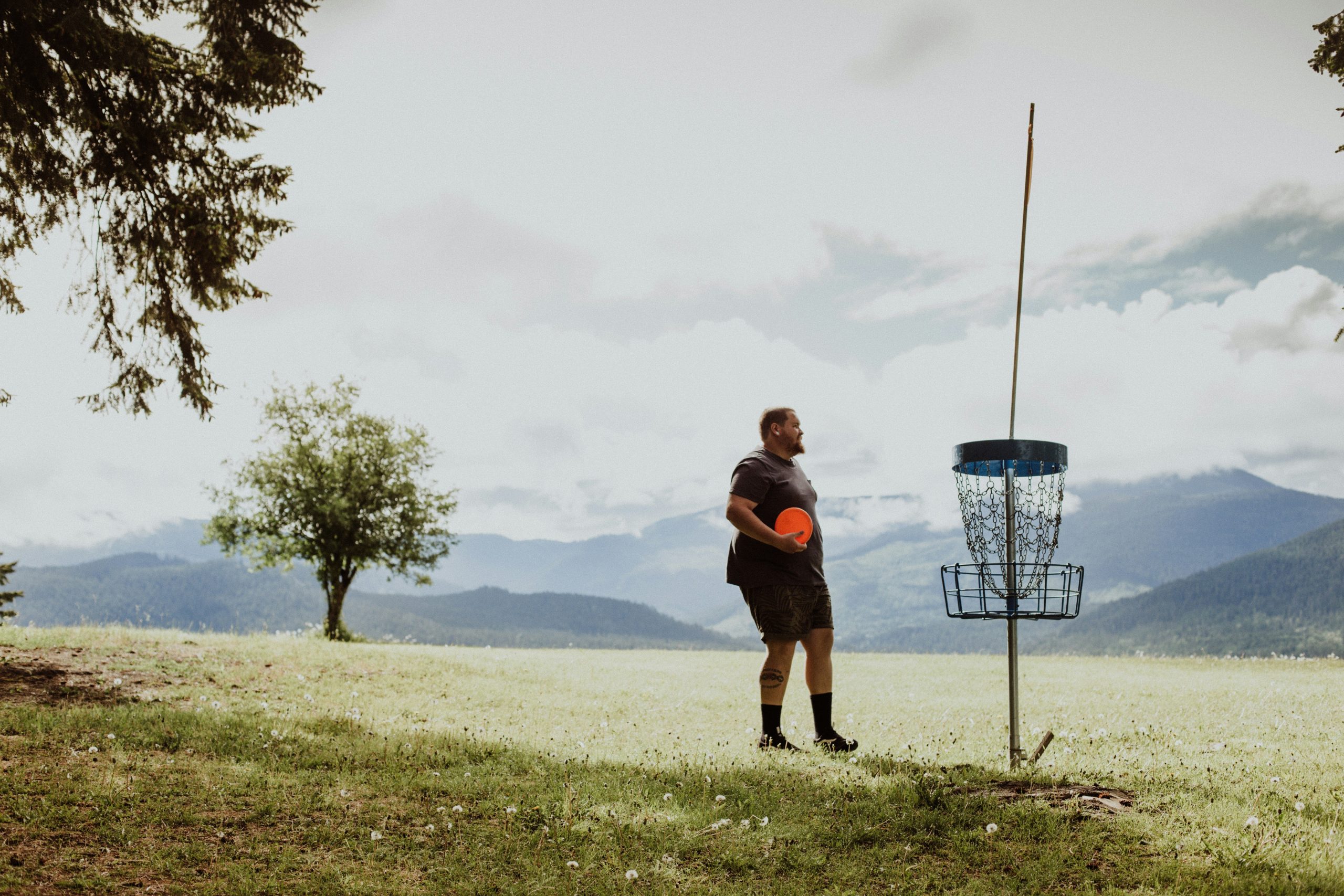 A man holding a plastic disc on a field beside a disc golf course basket target. A man holding a plastic disc on a field beside a disc golf course basket target.