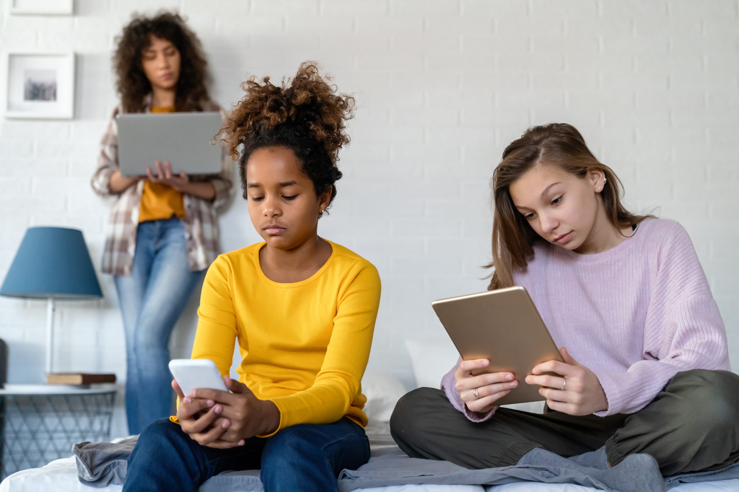 Des adolescentes assises sur un lit utilisent un téléphone, une tablette et un ordinateur portable.   Des adolescentes assises sur un lit utilisent un téléphone, une tablette et un ordinateur portable.